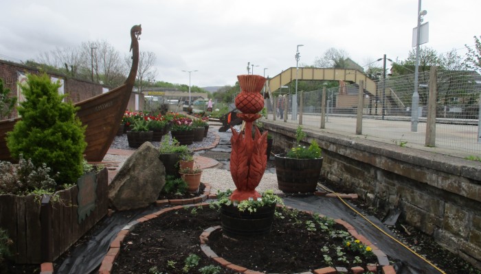 Plants, a wooden thistle statue and the bow of Viking long boat in Largs station community garden