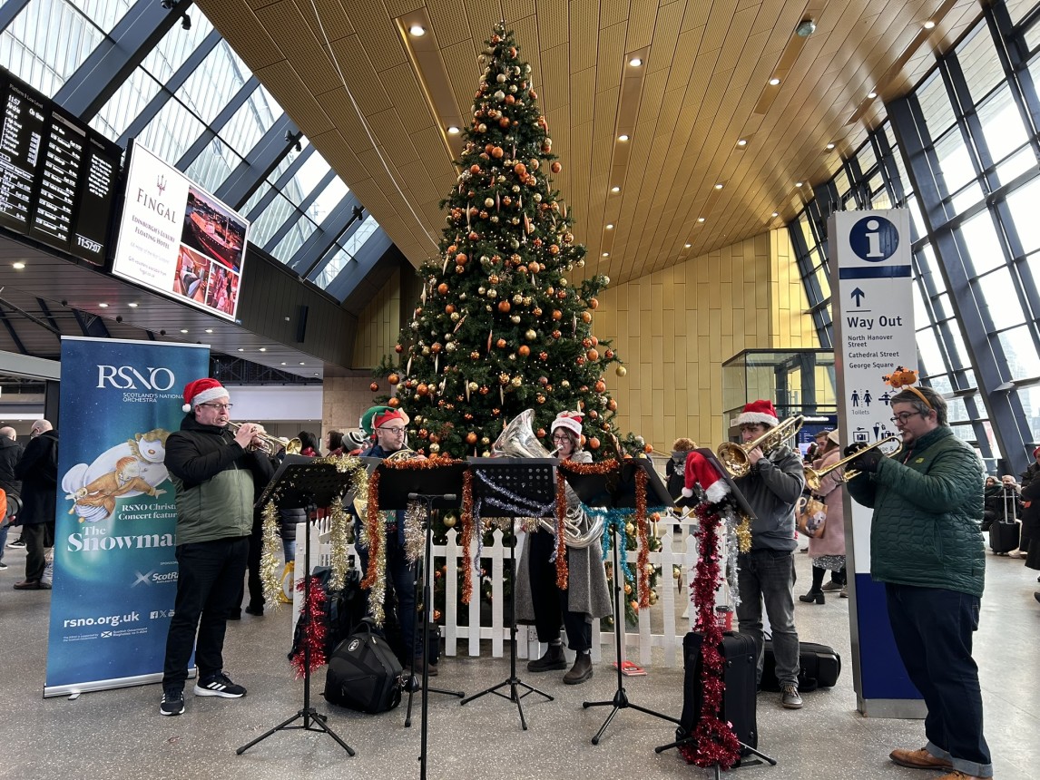 A group of brass musicians from the RSNO play carols in front of a Christmas Tree on Glasgow Queen St station concourse.