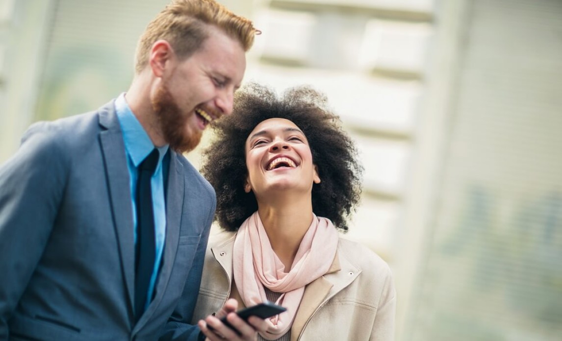Male and female laughing together, wearing smart attire, while man holds mobile phone