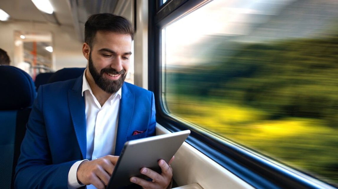 Business man travelling on a train with a tablet in his hand and the scenery from train window blurred behind him. 